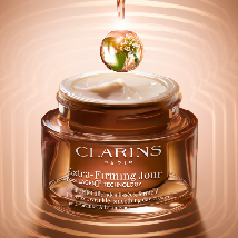Clarins Extra-Firming Jour Tagescreme in einem bronzefarbenen Glastiegel mit weißem Deckel. Darüber schwebt ein grün-goldener Tropfen mit Blattstruktur. Der Hintergrund ist warm beige mit radialen Lichtkreisen.