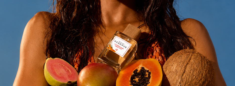 Frau hält Clean Reserve Sun-Kissed Papaya Parfum vor Früchten wie Guave, Mango und Papaya
