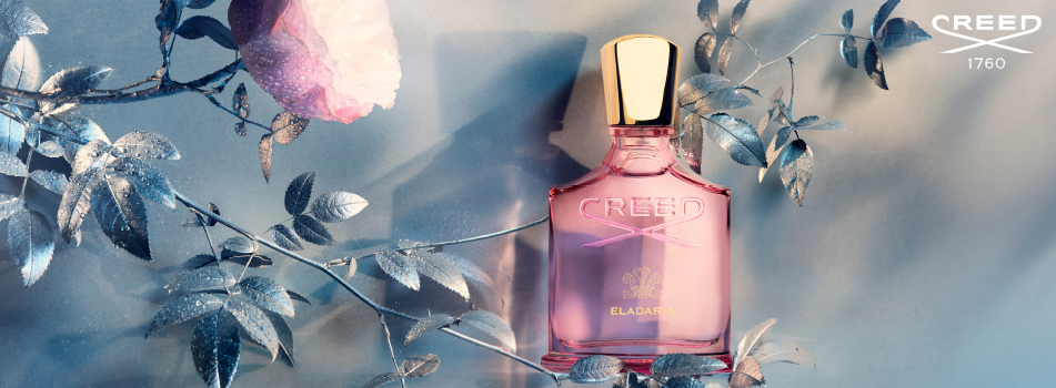 Creed Eladaria Parfumflakon in zartem Rosa mit goldenem Verschluss. Der Flakon steht vor einem pastellfarbenen Hintergrund mit schimmernden, silberblauen Blättern und einer halb geöffneten Rose. Oben rechts das Creed-Logo mit der Jahreszahl 1760.
