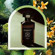 Dunkler Parfumflakon „Le Couvent Figu“ steht im Licht, umgeben von grünen Blättern und gelb-orangen Blüten, die an Feigenbäume erinnern.