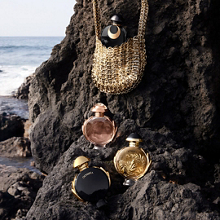 Olympéa Parfumkollektion von Rabanne mit mehreren Flakons auf dunklen Felsen am Meer, ergänzt durch eine goldene Tasche.