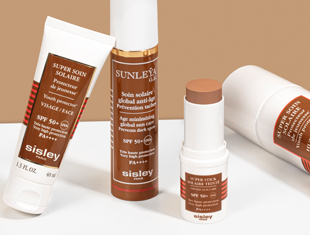 Vier Sonnenschutzprodukte von Sisley Paris auf heller Fläche vor beigefarbenem Hintergrund: eine Tube, ein Pumpspender, ein Stick mit herausgedrehtem Produkt und ein liegender Zylinder. Alle Produkte sind weiß mit braunen Etiketten und tragen Aufschriften wie 'Super Soin Solaire' oder 'Sunleÿa', sowie Hinweise auf SPF 50+ Sonnenschutz.